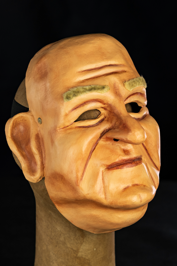 A máscara representa Bubi, um homem branco no início da velhice que possui um rosto grande e arredondado. Possui uma cor de pele clara e levemente avermelhada por todo o trabalho na terra que realiza debaixo de sol. Seu nariz é um típico “nariz batata” que revela sua ascendência germânica. Apesar de tudo preserva uma expressão quase calma, como se estivesse escutando alguém lhe contar algo, ou rindo baixinho de alguma piada contada por alguém, Seu queixo possui um vinco que o divide no meio e a máscara possui uma par de grandes orelhas e a aplicação de pelos sintéticos na região da sobrancelha.