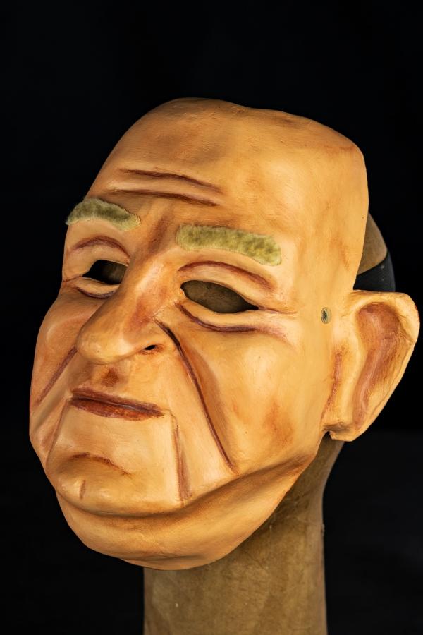 A máscara representa Bubi, um homem branco no início da velhice que possui um rosto grande e arredondado. Possui uma cor de pele clara e levemente avermelhada por todo o trabalho na terra que realiza debaixo de sol. Seu nariz é um típico “nariz batata” que revela sua ascendência germânica. Apesar de tudo preserva uma expressão quase calma, como se estivesse escutando alguém lhe contar algo, ou rindo baixinho de alguma piada contada por alguém, Seu queixo possui um vinco que o divide no meio e a máscara possui uma par de grandes orelhas e a aplicação de pelos sintéticos na região da sobrancelha.