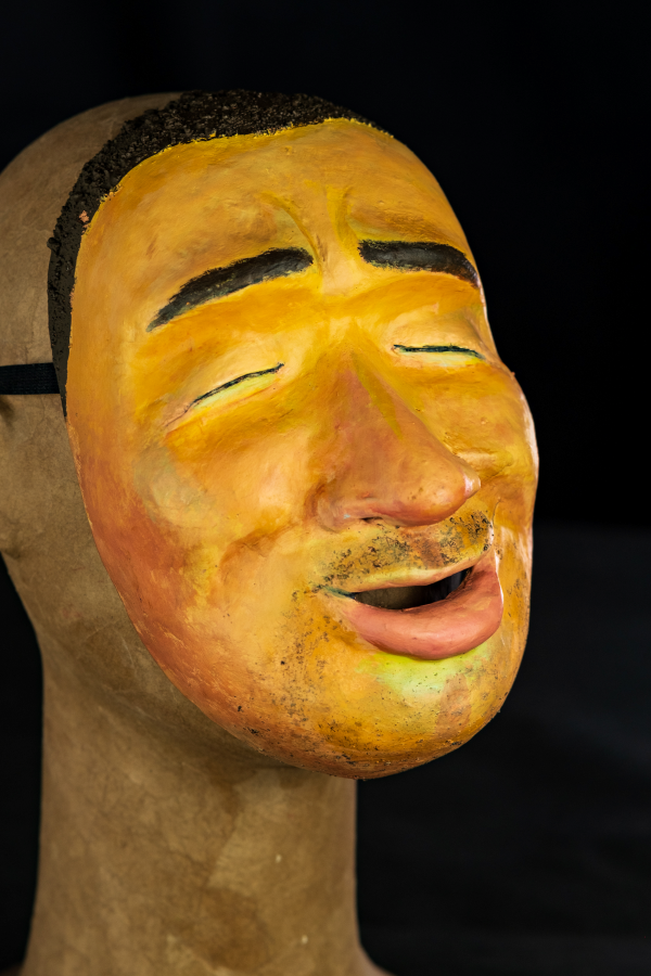 De olhos fechados, como quem olha para dentro de si, a máscara tem muitos detalhes. Um nariz grande, cobre o meio da face e tem sua ponta avermelhada. A boca está levemente aberta e tem a sombra de um bigode e uma barba por crescer. Sua pele é branca, com detalhes em vermelho e azul pelo rosto. A expressão é de calmaria.