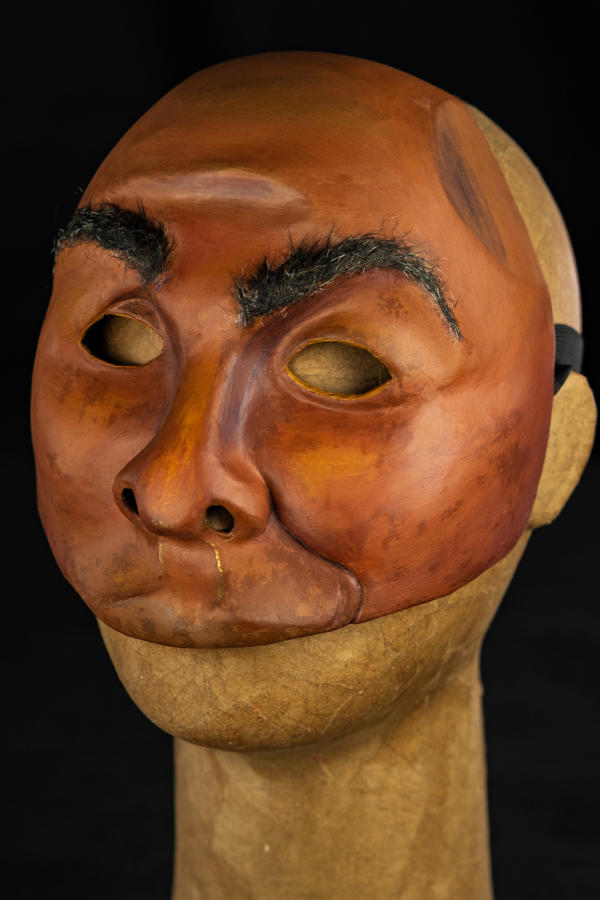 Na foto um rosto assimétrico com sobrancelhas arqueadas e lábios carnudos curvados para a esquerda. Sua pele é negra, com tonalidades roxas, amareladas e contornos dourados. O nariz é grande, mas achatado. Tem os olhos vazados, em contornos simples e simétricos. As bochechas são pequenas, características de um rosto firme. A expressão é de desconfiança e deboche, como quem escuta um causo muito aumentado.