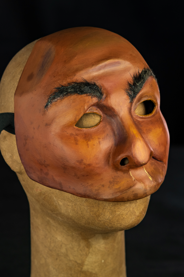 Na foto um rosto assimétrico com sobrancelhas arqueadas e lábios carnudos curvados para a esquerda. Sua pele é negra, com tonalidades roxas, amareladas e contornos dourados. O nariz é grande, mas achatado. Tem os olhos vazados, em contornos simples e simétricos. As bochechas são pequenas, características de um rosto firme. A expressão é de desconfiança e deboche, como quem escuta um causo muito aumentado.