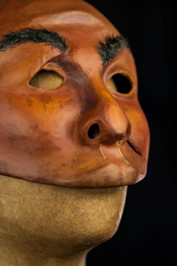 Na foto um rosto assimétrico com sobrancelhas arqueadas e lábios carnudos curvados para a esquerda. Sua pele é negra, com tonalidades roxas, amareladas e contornos dourados. O nariz é grande, mas achatado. Tem os olhos vazados, em contornos simples e simétricos. As bochechas são pequenas, características de um rosto firme. A expressão é de desconfiança e deboche, como quem escuta um causo muito aumentado.