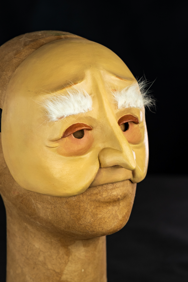 Uma meia máscara, branco-amarelada, com sobrancelhas longas, brancas e macias. Expressa um leve sorriso em sua cara de nariz longo e franzino. Os olhos são cansados acompanhados de olheiras profundas de quem já viveu muito.