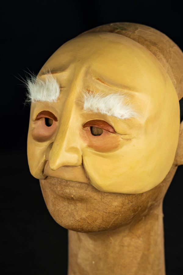 Uma meia máscara, branco-amarelada, com sobrancelhas longas, brancas e macias. Expressa um leve sorriso em sua cara de nariz longo e franzino. Os olhos são cansados acompanhados de olheiras profundas de quem já viveu muito.