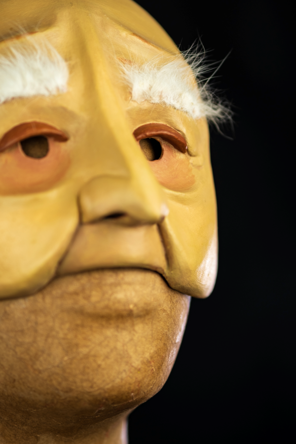 Uma meia máscara, branco-amarelada, com sobrancelhas longas, brancas e macias. Expressa um leve sorriso em sua cara de nariz longo e franzino. Os olhos são cansados acompanhados de olheiras profundas de quem já viveu muito.