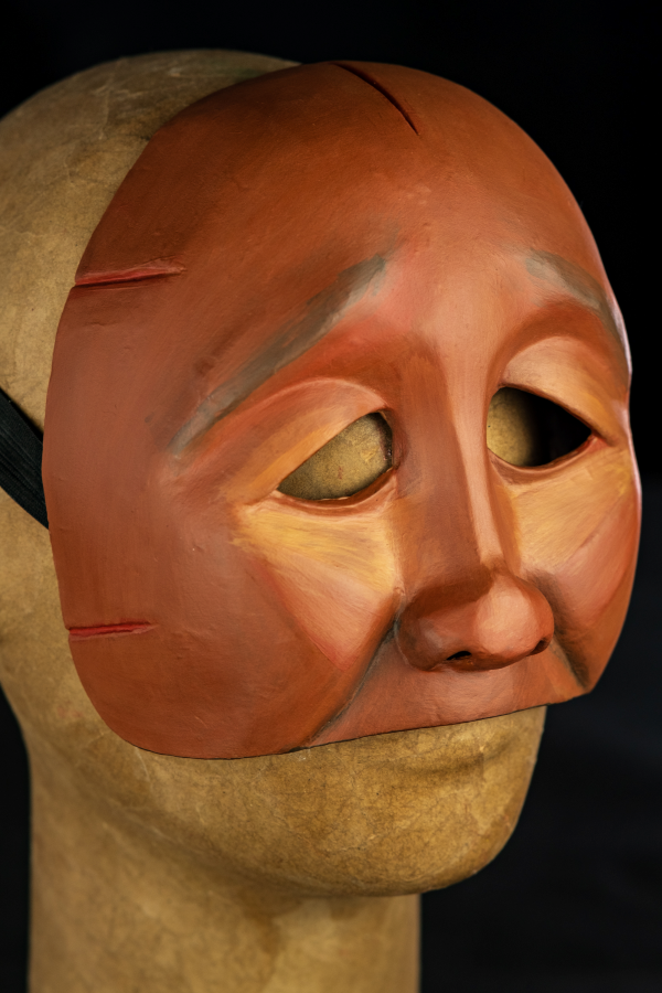 Um rosto pequeno, de traços simples, sem boca ou queixo. A pele é de um tom marrom avermelhado que revela sua descendência negra e indígena. Os olhos são pequenos e o olhar caído com olhos vazados em linhas simples. As bochechas são pequenas e o rosto é magro. Em toda as formas, uma estética diamantada toma conta desse rosto, que parece marcado por algo ou alguém. Há uma expressão de cansaço ou dor nesta cara incompleta. Há também detalhes nas laterais do rosto como um vinco. Vermelho e verde também são cores que complementam este objeto.