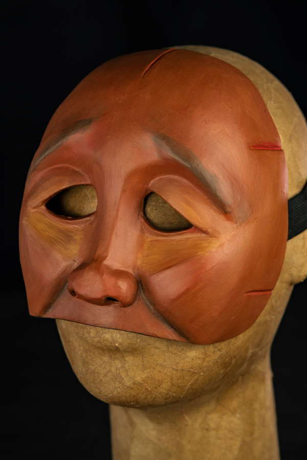 Um rosto pequeno, de traços simples, sem boca ou queixo. A pele é de um tom marrom avermelhado que revela sua descendência negra e indígena. Os olhos são pequenos e o olhar caído com olhos vazados em linhas simples. As bochechas são pequenas e o rosto é magro. Em toda as formas, uma estética diamantada toma conta desse rosto, que parece marcado por algo ou alguém. Há uma expressão de cansaço ou dor nesta cara incompleta. Há também detalhes nas laterais do rosto como um vinco. Vermelho e verde também são cores que complementam este objeto.