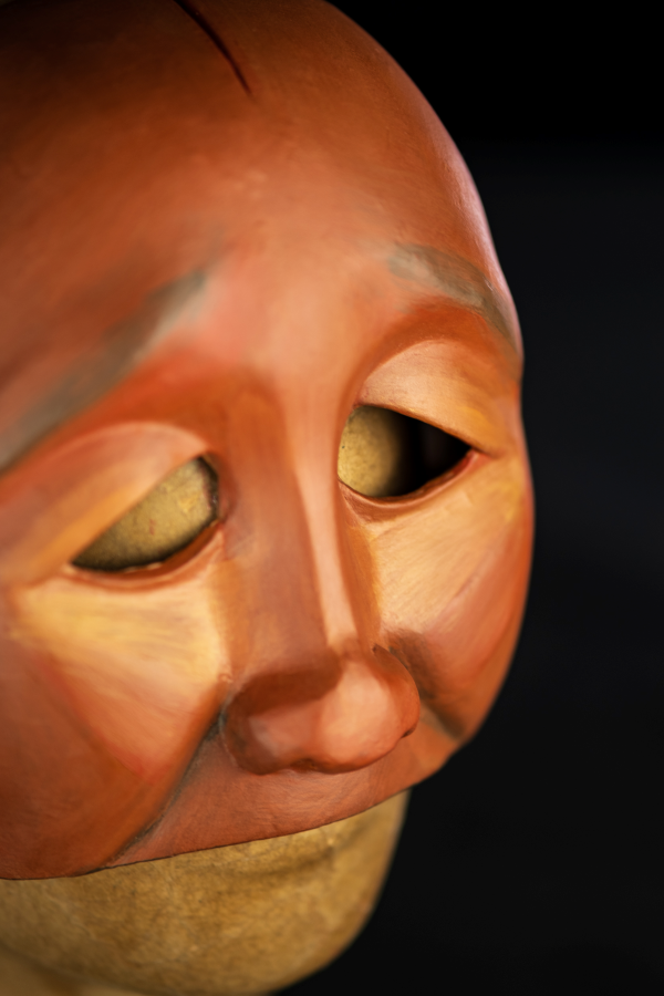 Um rosto pequeno, de traços simples, sem boca ou queixo. A pele é de um tom marrom avermelhado que revela sua descendência negra e indígena. Os olhos são pequenos e o olhar caído com olhos vazados em linhas simples. As bochechas são pequenas e o rosto é magro. Em toda as formas, uma estética diamantada toma conta desse rosto, que parece marcado por algo ou alguém. Há uma expressão de cansaço ou dor nesta cara incompleta. Há também detalhes nas laterais do rosto como um vinco. Vermelho e verde também são cores que complementam este objeto.