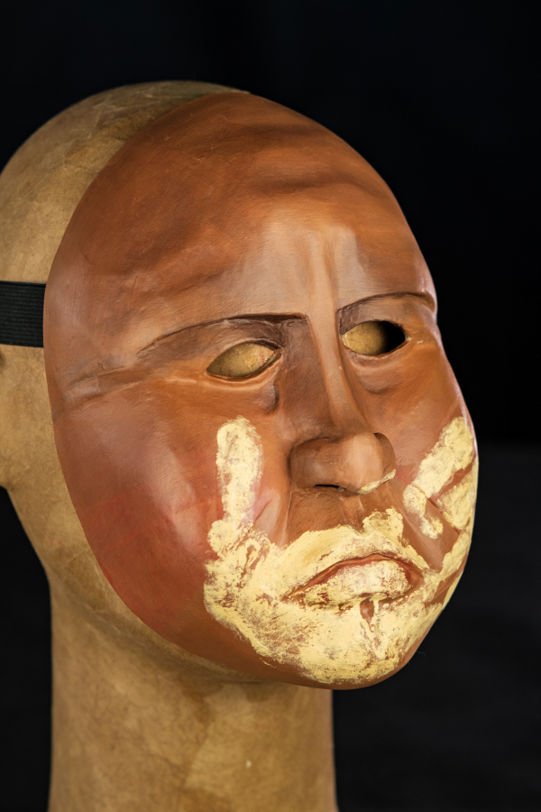 Uma máscara inteira, carrancuda, de olhar muito severo e fundo. Sobrancelhas baixas, enfatizando sua bravura. Rosto cheio de marcas que precisam ser sentidas nas pontas dos dedos, pois são registros da sua identidade e história. Sobre sua boca fechada, as marcas de uma mão que o silencia. Uma mão, feita do barro do rio que lhe serviu de morada.