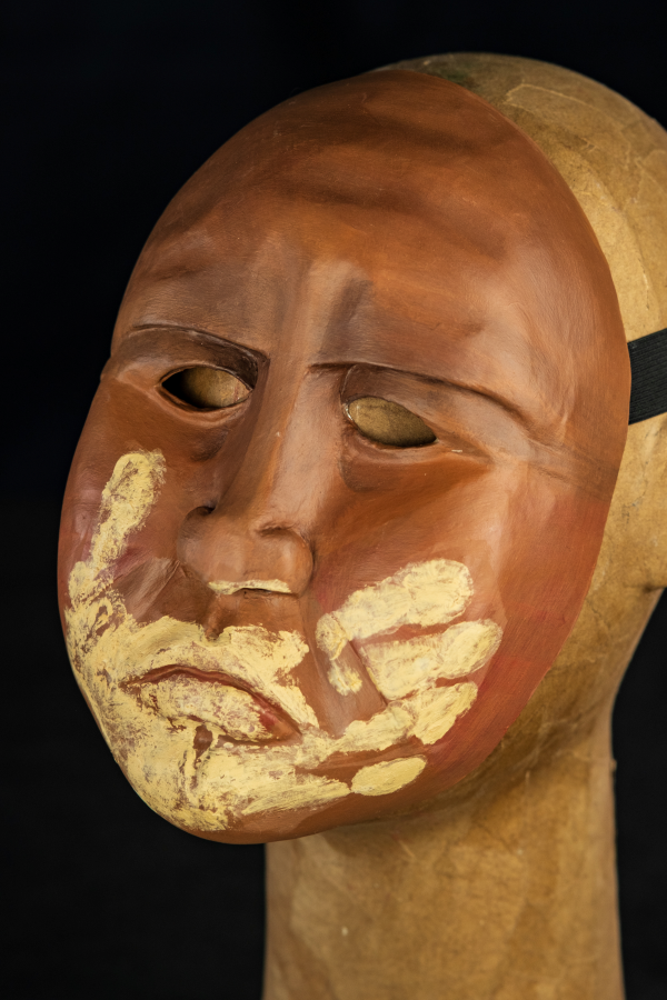 Uma máscara inteira, carrancuda, de olhar muito severo e fundo. Sobrancelhas baixas, enfatizando sua bravura. Rosto cheio de marcas que precisam ser sentidas nas pontas dos dedos, pois são registros da sua identidade e história. Sobre sua boca fechada, as marcas de uma mão que o silencia. Uma mão, feita do barro do rio que lhe serviu de morada.