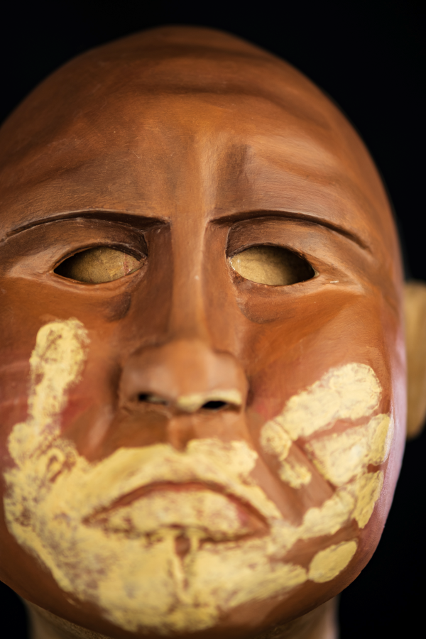 Uma máscara inteira, carrancuda, de olhar muito severo e fundo. Sobrancelhas baixas, enfatizando sua bravura. Rosto cheio de marcas que precisam ser sentidas nas pontas dos dedos, pois são registros da sua identidade e história. Sobre sua boca fechada, as marcas de uma mão que o silencia. Uma mão, feita do barro do rio que lhe serviu de morada.